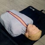 Manekin do resuscytacji leży na czarnej macie na podłodze. Ma beżową głowę i szarą pokrowiec. Tło to jasne panele.