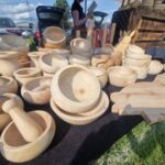 Na zdjęciu widać drewniane naczynia i moździerze na stoisku. Kolory są naturalne, beżowe, w tle samochody. Tło zielone, trawa i niebo.