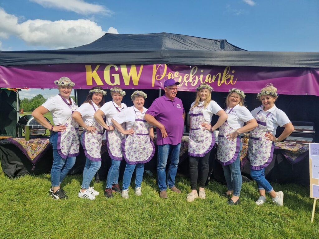 Grupa ludzi w białych koszulkach i fioletowych fartuchach stoi przed namiotem z napisem KGW Porębianki. Tło to zielona trawa i niebo z chmurami. Osoby mają na głowach kwiatowe wianki.