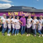 Grupa ludzi w białych koszulkach i fioletowych fartuchach stoi przed namiotem z napisem KGW Porębianki. Tło to zielona trawa i niebo z chmurami. Osoby mają na głowach kwiatowe wianki.