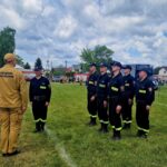 Strażacy w mundurach ćwiczą na zielonym polu. Niebieskie niebo z chmurami w tle. Wokół zielone drzewa i budynki.