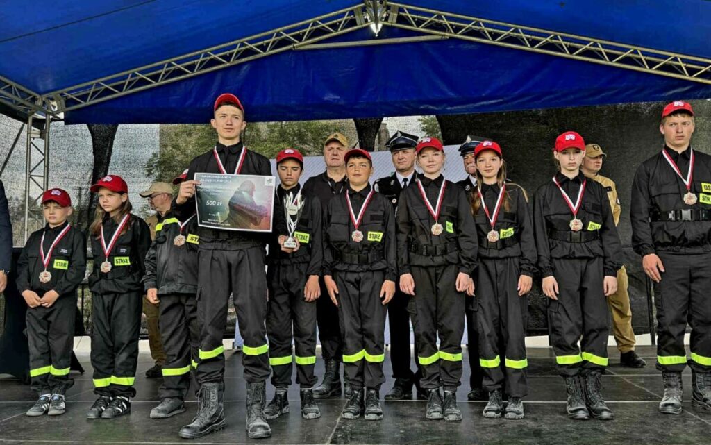 Grupa młodych strażaków w czarnych uniformach i czerwonych czapkach stoi na scenie pod niebieskim namiotem, prezentując medale i dyplom.