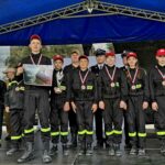 Grupa młodych strażaków w czarnych uniformach i czerwonych czapkach stoi na scenie pod niebieskim namiotem, prezentując medale i dyplom.