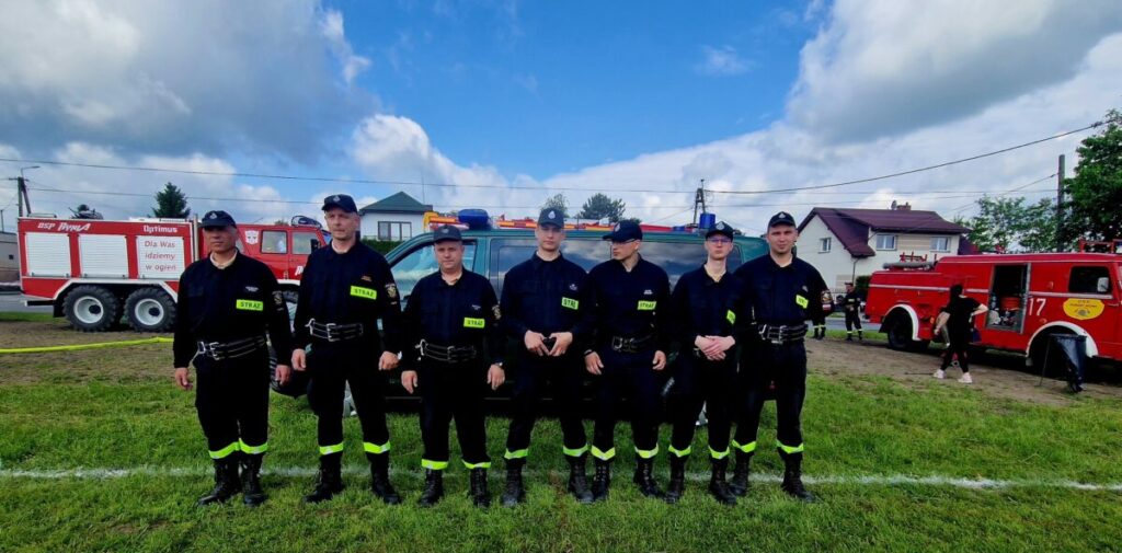Grupa strażaków w ciemnych uniformach stoi na trawie. W tle dwa czerwone wozy strażackie i domy pod błękitnym niebem z chmurami.