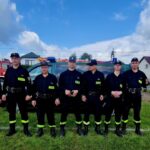 Grupa strażaków w ciemnych uniformach stoi na trawie. W tle dwa czerwone wozy strażackie i domy pod błękitnym niebem z chmurami.