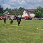 Strażacy ćwiczą na zielonej łące z żółtymi wężami strażackimi. W tle czerwone wozy strażackie i białe domy. Na niebie białe chmury.