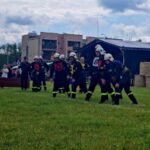 Strażacy w strojach ochronnych przygotowują się do zawodów na trawie. W tle widać namiot, budynki i chmury na niebie. Po prawej stronie są ustawione bele siana.