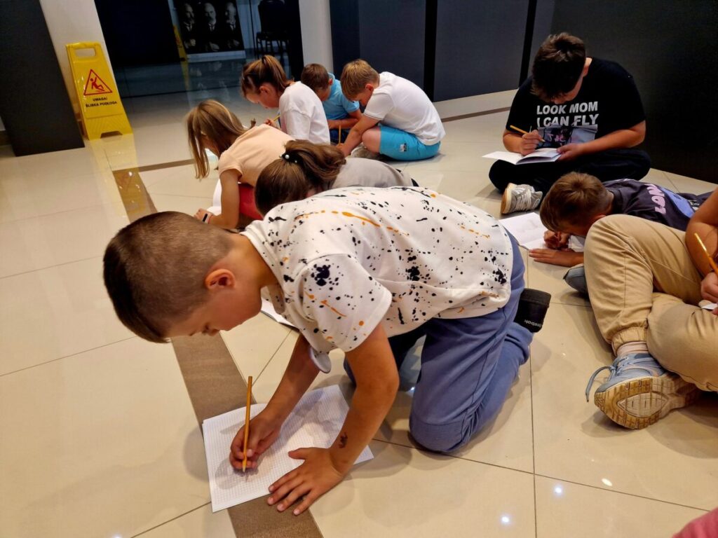 Grupa dzieci siedzi na jasnej podłodze, pisząc na kartkach papieru. Tło jest ciemne, z żółtym znakiem ostrzegawczym. Dzieci mają kolorowe ubrania.