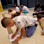 Grupa dzieci siedzi na jasnej podłodze, pisząc na kartkach papieru. Tło jest ciemne, z żółtym znakiem ostrzegawczym. Dzieci mają kolorowe ubrania.