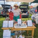 Dwójka ludzi przy stoisku z naturalnymi produktami na tle otwartych bagażników samochodów. Jasne kolory przy ciastkach na drewnianym stole. Namiot i zielona trawa nadają atmosferę pikniku.