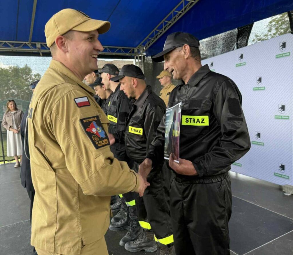 Strażacy w czarnych mundurach i żółtych naszywkach STRAŻ otrzymują gratulacje. Tło to niebieski namiot i biała ścianka z logo gminy. Atmosfera jest uroczysta.