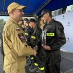 Strażacy w czarnych mundurach i żółtych naszywkach STRAŻ otrzymują gratulacje. Tło to niebieski namiot i biała ścianka z logo gminy. Atmosfera jest uroczysta.