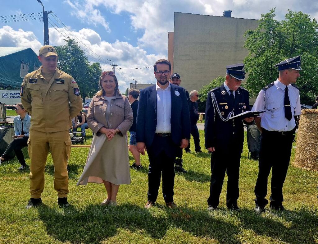 Grupa pięciu osób stoi na tle zielonej trawy. Trzy osoby w mundurach, jedna w eleganckiej sukience. Tło tworzą drzewa, niebo i budynek.