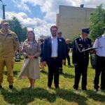 Grupa pięciu osób stoi na tle zielonej trawy. Trzy osoby w mundurach, jedna w eleganckiej sukience. Tło tworzą drzewa, niebo i budynek.