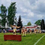 Strażak w żółtym mundurze skacze przez przeszkodę na trawie. W tle kilku strażaków, budynki i drzewa. Niebo zachmurzone.