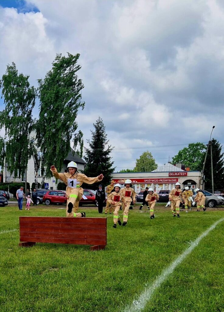 Strażak w żółtym mundurze skacze przez przeszkodę na trawie. W tle kilku strażaków, budynki i drzewa. Niebo zachmurzone.