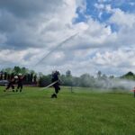 Strażacy w hełmach ćwiczą z wężami na zielonym polu. W tle niebieskie niebo z chmurami. Z boku stoi dom i drzewa.