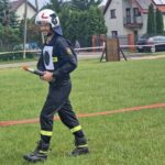 Mężczyzna w stroju strażaka z uśmiechem trzyma narzędzie na trawie. W tle domy i zaparkowane auta. Dzień, zielona trawa.