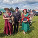 Trzy osoby w stylowych strojach na tle zielonej trawy i nieba z chmurami. Kobieta po lewej w bordowej sukni i kapeluszu, mężczyzna w garniturze z melonikiem, kobieta po prawej w zielonej sukni z czerwonym kwiatowym