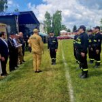 Grupa strażaków w czarnych mundurach stoi na trawie naprzeciw dwóch osób. Obecni są także inni w cywilnych ubraniach. W tle namiot, drzewa i niebo z chmurami.