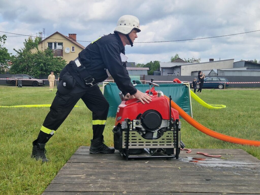 Na zdjęciu strażak w czarnym mundurze obsługuje czerwoną pompę wodną na trawie. W tle widoczne są domy i samochody. Niebo jest pochmurne.