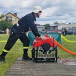 Na zdjęciu strażak w czarnym mundurze obsługuje czerwoną pompę wodną na trawie. W tle widoczne są domy i samochody. Niebo jest pochmurne.