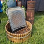 Drewniana tara leży na beczce na trawie, obok niej niebieski wazon i drewniane naczynia. W tle czarny stół i kolorowy bieżnik.