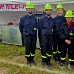 Czwórka osób w czarnych mundurach i zielonych hełmach pozuje na tle białego namiotu. Tło zawiera napis OSP MLECIN. Trawa pod stopami jest zielona i gęsta.