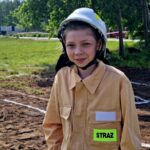 Na zdjęciu młoda osoba w beżowym stroju strażaka z białym kaskiem. Tło to zielona trawa i drzewa. Na kombinezonie widoczny jest napis STRAŻ.