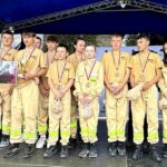 Grupa młodzieży w mundurach strażackich stoi na scenie, trzymając medale. Obok dorosły w garniturze. Tło w kolorze niebieskim.