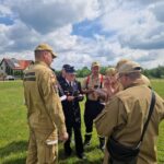 Grupa strażaków na zielonym polu, ubrani w jasne mundury pod pochmurnym niebem. W tle domy i drzewa. Trwa narada.