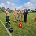 Grupa osób w beżowych i czarnych ubraniach stoi na zielonym boisku. Na trawie widać czerwony sprzęt strażacki. W tle są wozy strażackie i niebo z chmurami.