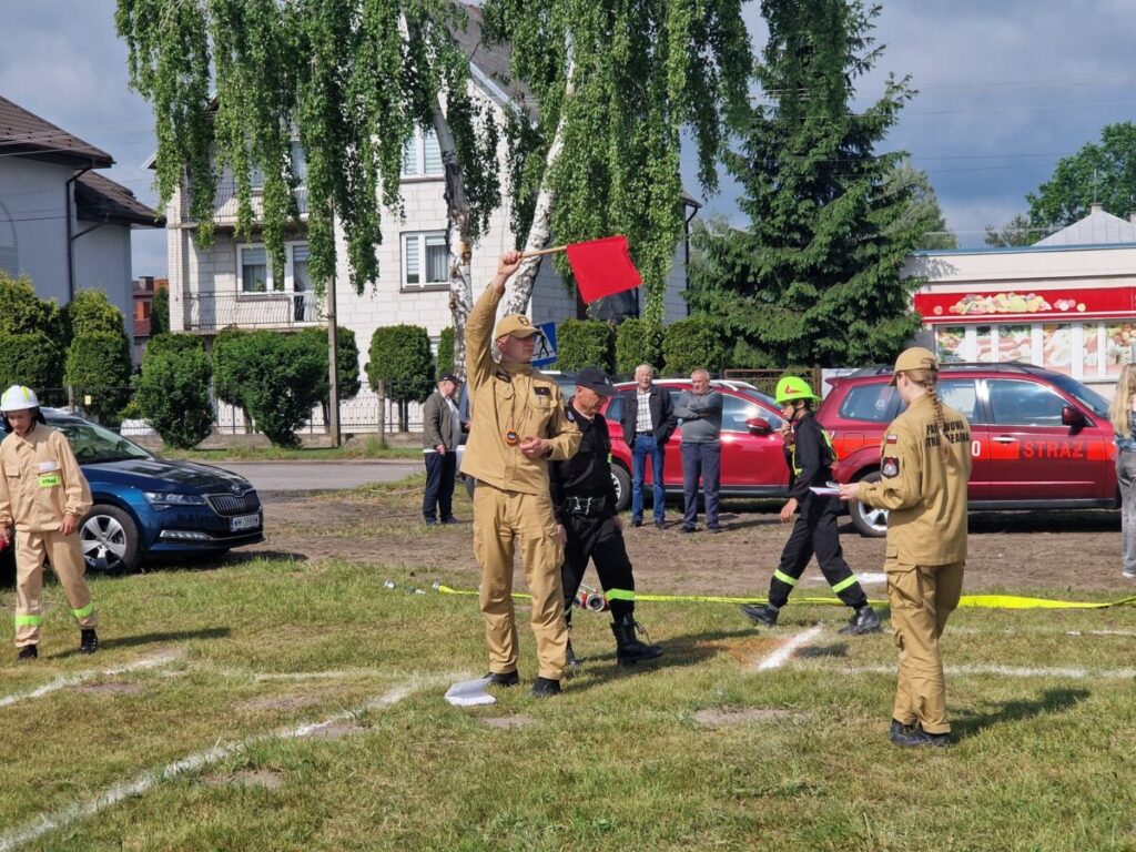 Na zielonej trawie stoją strażacy w beżowych mundurach z czerwonymi flagami. W tle biały budynek i czerwone auta strażackie. Dzień jest słoneczny.
