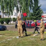 Na zielonej trawie stoją strażacy w beżowych mundurach z czerwonymi flagami. W tle biały budynek i czerwone auta strażackie. Dzień jest słoneczny.