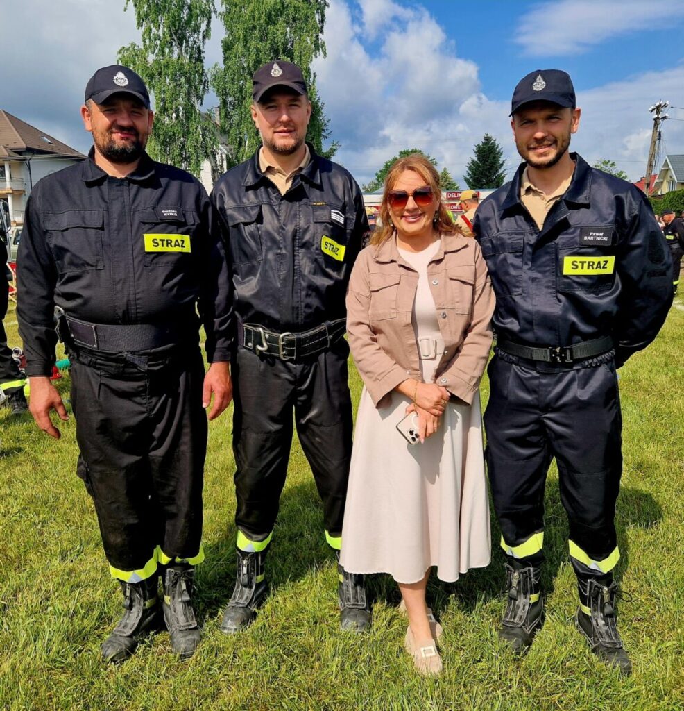 Czterech ludzi stoi na trawie. Trzech mężczyzn w strażackich mundurach, kobieta w jasnej sukience. Tło to budynki i drzewa.