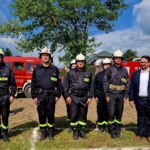 Grupa strażaków w mundurach stoi na trawie z czerwonymi wozami strażackimi w tle. Obok nich, mężczyzna w garniturze. Na niebie widać chmury i drzewa.