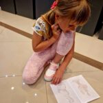 Dziewczynka w kolorowym ubraniu rysuje na podłodze. Ma różowe spodnie i trzyma ołówek. Na ziemi leży otwarty zeszyt.