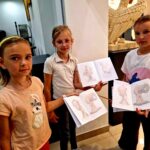 Trzy dziewczynki z zeszytami pokazującymi rzeźby. Stoją w jasnym muzeum z rzeźbą w tle. Ubrane w kolorowe koszulki.