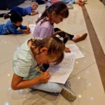 Dzieci siedzą na jasnej podłodze, rysując w zeszytach. Mają na sobie kolorowe ubrania. W tle inni również rysują.