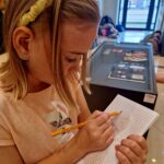 Dziewczynka rysuje ołówkiem na kratkowanym papierze, nosząc żółtą opaskę. Ubrana w beżową bluzkę. W tle są przeszklone drzwi i wystawa.