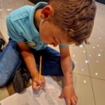 Chłopiec w niebieskiej koszulce rysuje na kartce w kratkę. Siedzi na jasnej podłodze z ołówkiem w ręku. Obok widać szkic i cienie.