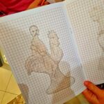 Praca na kartce w kratkę, pokazuje naszkicowany ołówkiem zarys koguta. W tle widać ciepłe, jasne oświetlenie. Dookoła kartki widać dłonie trzymające ołówek.