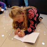 Dziewczynka na podłodze rysuje ołówkiem w zeszycie. Ma różową bluzę, wokół jasne kafelki. W tle inni zajęci pisaniem.