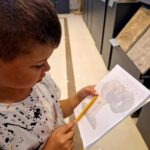 Chłopiec w muzeum rysuje rzeźbę ołówkiem na kartce w kratkę. Tło jasne, szare rzeźby na wystawie. Ma kolorową koszulkę.