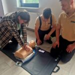 Na zdjęciu osoba wykonuje resuscytację na fantomie, dwie inne osoby patrzą. Tło to jasny pokój z oknem. Kolory stonowane, dominują beże i brązy.