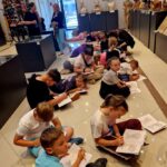 Grupa dzieci siedzi na podłodze w muzeum, rysując z notatnikami. W tle stoją popiersia i obrazy, ściany są białe. Po lewej stronie są kolorowe dekoracje.