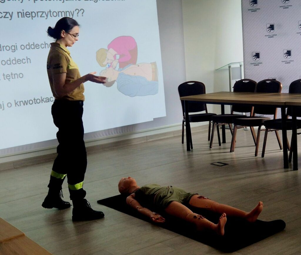 Kobieta w mundurze uczy pierwszej pomocy. Na podłodze leży manekin. W tle rzutnik z instrukcją resuscytacji.