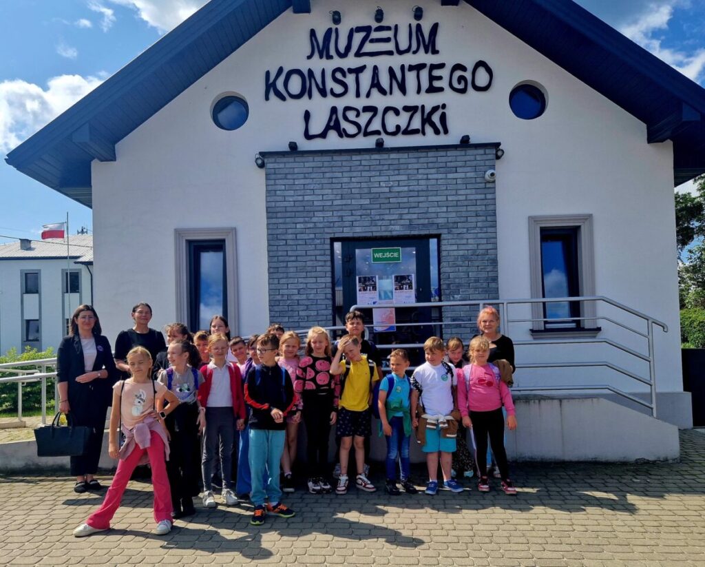 Grupa dzieci i dorosłych stoi przed białym budynkiem z napisem Muzeum Konstantego Laszczki. Budynek ma szare elementy i niebieski dach. W tle widać biało-czerwony dach innego budynku.