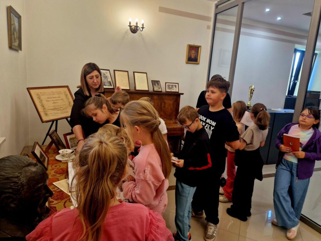 Na zdjęciu grupa dzieci w muzeum zwiedza ekspozycję z przewodniczką. Dzieci są skupione wokół stołu z eksponatami. Tło stanowią obrazy i certyfikaty na ścianach.
