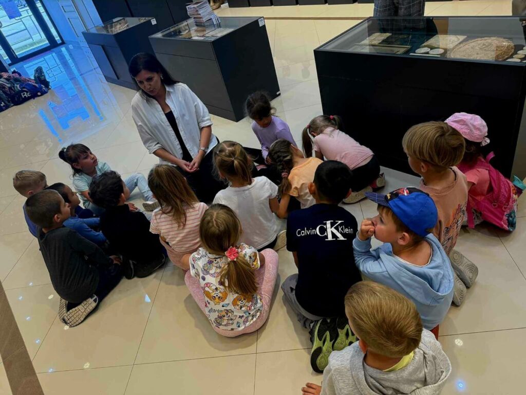 Grupa dzieci siedzi na jasnej podłodze wokół kobiety w białej koszuli. Tło stanowią ciemne gabloty muzealne. Dzieci są skupione i różnorodnie ubrane.